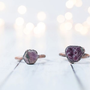 May include: Two copper rings with raw pink and purple gemstone settings. The rings are on a white wooden surface.