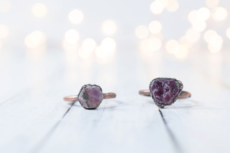 May include: Two copper rings with raw pink and purple gemstone settings. The rings are on a white wooden surface.