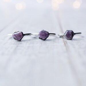 May include: Three silver rings with purple gemstone accents. The rings are arranged in a row on a white wooden surface.