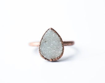 Op de afbeelding: Een ring in de vorm van een traan met een witte, getextureerde steen gezet in een koperkleurige band.