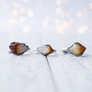 May include: Three silver rings with raw, natural, amber-colored gemstones. The rings are set on a white wooden surface.
