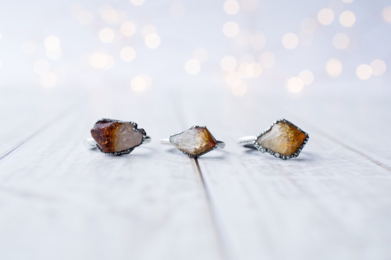 May include: Three silver rings with raw, natural, amber-colored gemstones. The rings are set on a white wooden surface.