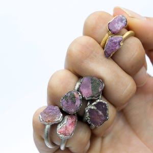 May include: A hand wearing several rings with pink and purple gemstone settings. The rings are made of silver and gold.