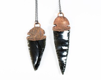 Puede incluir: Dos colgantes de punta de flecha de obsidiana negra con detalles en cobre en cadenas de plata. Las puntas de flecha son de diferentes tamaños y tienen un acabado oscuro y brillante.