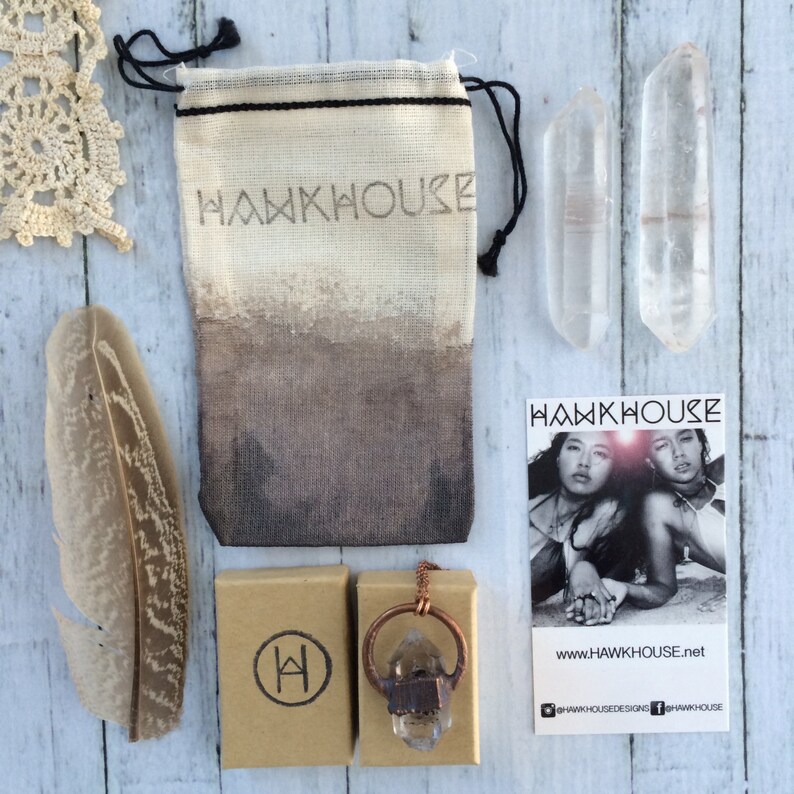 May include: A crystal pendant necklace with a copper chain and a raw crystal stone. The necklace is displayed on a brown box with the letter "H" in a circle. The necklace is packaged in a small white drawstring bag with the word "HAWKHOUSE" printed on it. Two clear quartz crystals are also shown in the image.