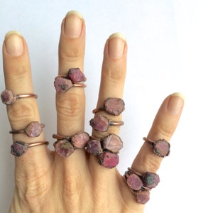 May include: A hand model wearing multiple rings with pink and red gemstone settings. The rings are made of copper wire and feature raw, uncut gemstones.