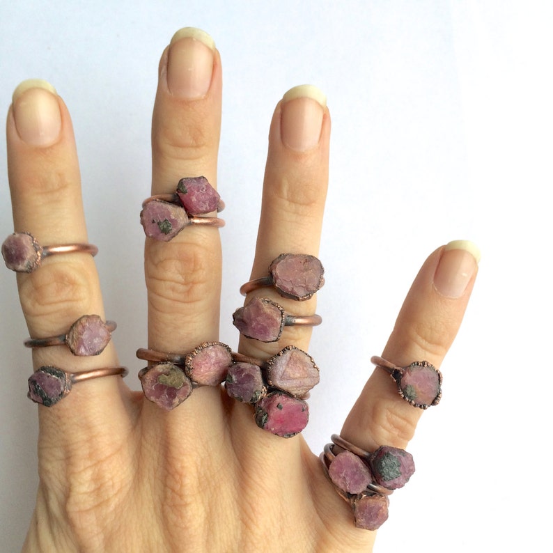 May include: A hand model wearing multiple rings with pink and red gemstone settings. The rings are made of copper wire and feature raw, uncut gemstones.