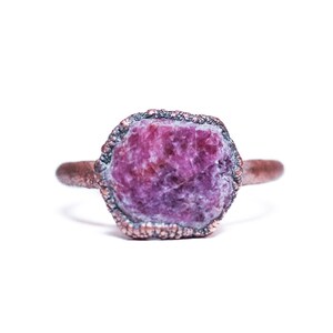 May include: A copper ring featuring a raw, pink ruby gemstone set in the centre. The ring has a textured band and a rough, natural look.