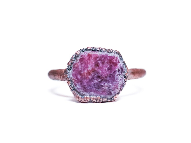 May include: A copper ring featuring a raw, pink ruby gemstone set in the centre. The ring has a textured band and a rough, natural look.