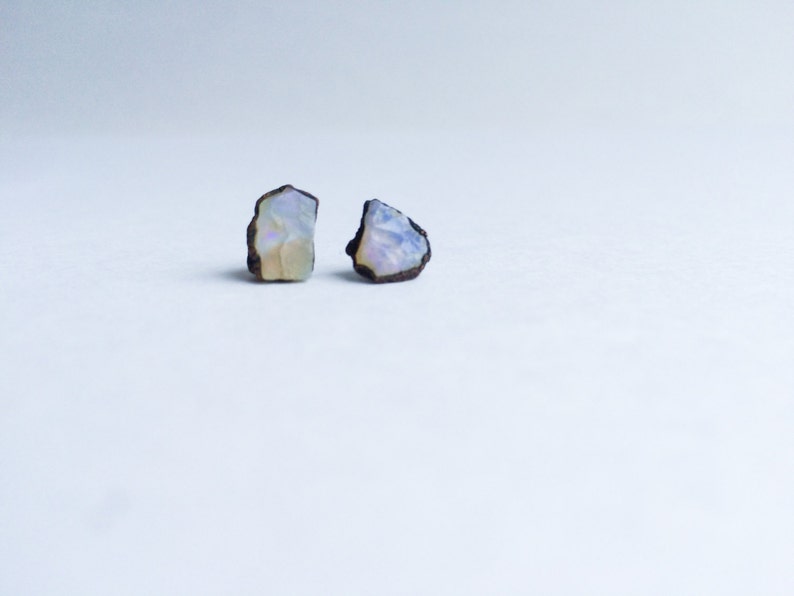 Pu&ograve; includere: Un paio di orecchini in opale iridescente con un aspetto grezzo e naturale. Gli orecchini sono incastonati in un metallo color bronzo.