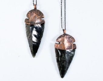 Puede incluir: Dos colgantes de punta de flecha de obsidiana con tapas de metal color cobre y cadenas oscuras. Las puntas de flecha son de color gris oscuro y tienen una superficie pulida y reflectante. Las tapas metálicas tienen un aspecto texturizado y martillado.