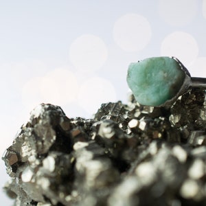 May include: A silver ring with a large, light green gemstone set in a rough, natural style. The ring is resting on a bed of small, metallic, gray crystals.