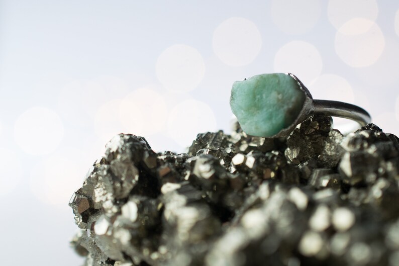 May include: A silver ring with a large, light green gemstone set in a rough, natural style. The ring is resting on a bed of small, metallic, gray crystals.