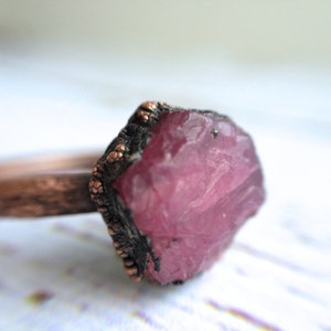 May include: A copper ring featuring a large, raw ruby gemstone set in the centre. The ring band is textured and has a hammered finish.