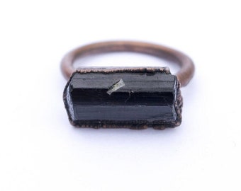 Puede incluir: Un anillo de turmalina negra con una banda de cobre. El anillo está engastado con una piedra de turmalina en bruto sin cortar.