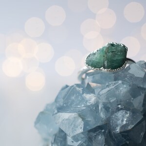 May include: A green emerald gemstone ring set in silver, resting on a bed of blue crystals. The ring features a raw, uncut emerald with a unique shape.