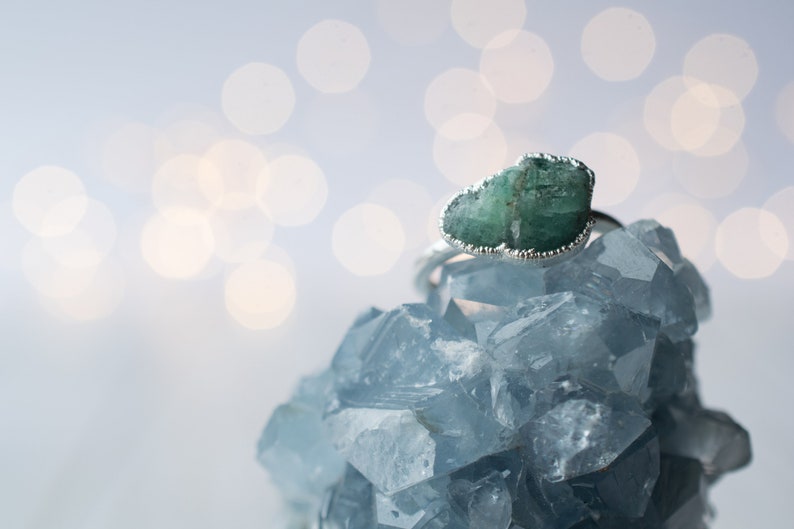 May include: A green emerald gemstone ring set in silver, resting on a bed of blue crystals. The ring features a raw, uncut emerald with a unique shape.