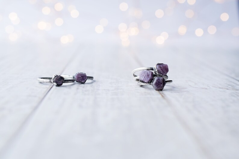 May include: Three silver rings with purple gemstone accents. The rings are stacked on a white wooden surface.