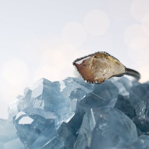 May include: A raw, yellow-brown crystal ring set in a silver band, resting on a bed of blue crystals.