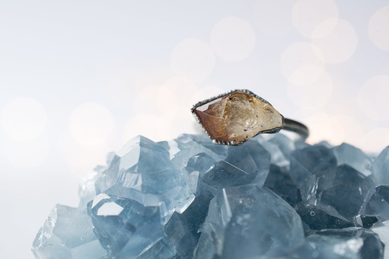 May include: A raw, yellow-brown crystal ring set in a silver band, resting on a bed of blue crystals.