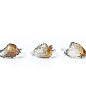 May include: Three silver rings with raw, irregular shaped, clear and yellow gemstones. The rings are set in a silver band with a textured edge.