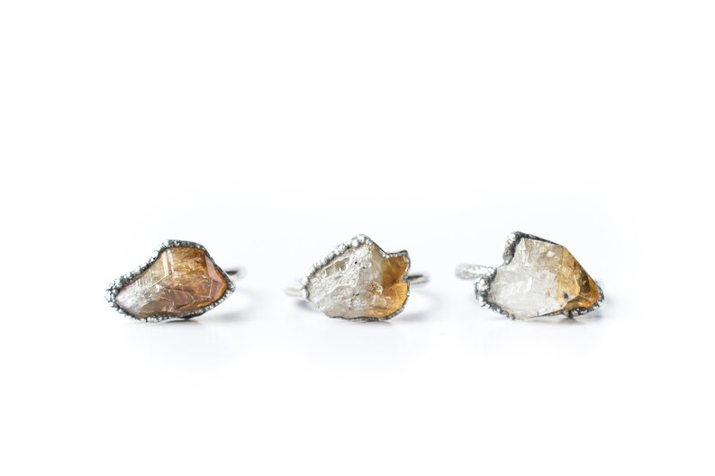 May include: Three silver rings with raw, irregular shaped, clear and yellow gemstones. The rings are set in a silver band with a textured edge.