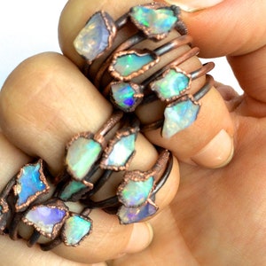 Puede incluir: Un conjunto de anillos envueltos en alambre de cobre con piedras preciosas de ópalo iridiscente. Los anillos están apilados uno encima del otro y están siendo sostenidos por una mano.