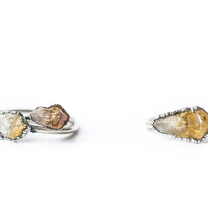 May include: Two silver rings with raw, uncut gemstones. One ring features a single, clear and yellow gemstone, while the other ring has two gemstones, one clear and one brown.