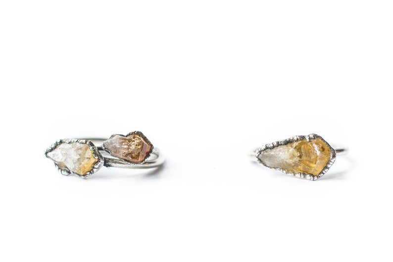May include: Two silver rings with raw, uncut gemstones. One ring features a single, clear and yellow gemstone, while the other ring has two gemstones, one clear and one brown.