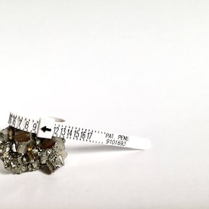 May include: White plastic ring sizer with black markings from 6 to 17. The sizer is resting on a pile of gray and silver pyrite crystals. The sizer has the text "PAT. PEND. 9101693" printed on it.
