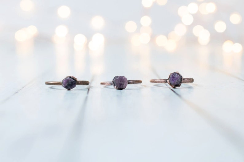 K&ouml;nnte beinhalten: Drei Kupferringe mit violetten Edelstein-Akzenten. Die Ringe sind in einer Reihe auf einer wei&szlig;en Holzoberfl&auml;che angeordnet.