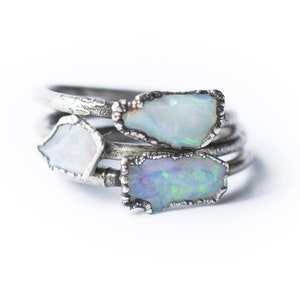 May include: Three silver rings with opal gemstones. The opals are a milky white with iridescent blue and green hues. The rings are stacked on top of each other.