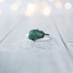 May include: A silver ring with a green gemstone set in the center. The ring is on a white wooden surface.