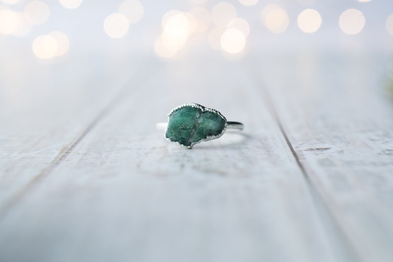 May include: A silver ring with a green gemstone set in the center. The ring is on a white wooden surface.
