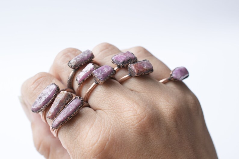 May include: A set of copper rings with pink and purple gemstone accents. The rings are stacked on a person's hand.
