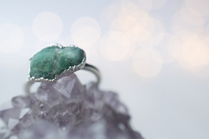 May include: A silver ring with a green gemstone set in a silver bezel. The ring is sitting on a bed of purple crystals.
