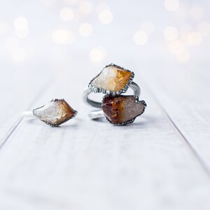 May include: Three silver rings with raw, natural, yellow and orange crystal stones. The rings are on a white wooden surface.