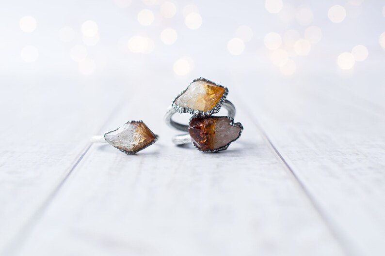 May include: Three silver rings with raw, natural, yellow and orange crystal stones. The rings are on a white wooden surface.