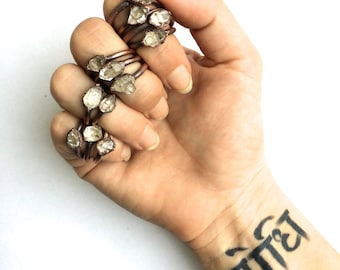 May include: A hand wearing multiple rings with raw quartz crystals set in copper wire. The rings are stacked on each finger. The hand has a tattoo on the wrist that says "बॉन्ड".