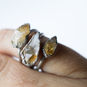 May include: Three silver rings with raw, natural, yellow and clear quartz stones. The rings are stacked on a finger.
