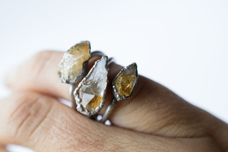 May include: Three silver rings with raw, natural, yellow and clear quartz stones. The rings are stacked on a finger.