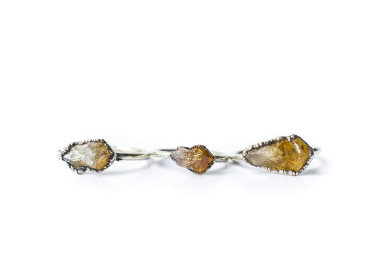May include: Three silver rings with raw crystal stones. The stones are a light yellow, a darker yellow, and a clear crystal.