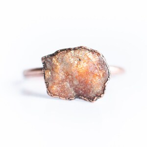 Puede incluir: Un anillo de cobre con una piedra de sol en bruto de forma irregular engastada en el centro. La piedra es una mezcla de naranja, marrón y blanco.