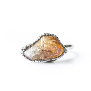 May include: A silver ring with a raw, irregular shaped, amber-colored gemstone set in the center.