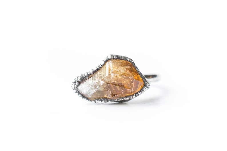 May include: A silver ring with a raw, irregular shaped, amber-colored gemstone set in the center.