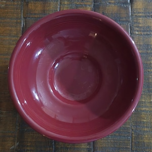 May include: A deep red ceramic bowl with a glossy finish. The bowl has a rounded shape with a wide rim and a slightly recessed center. The color is a rich, solid hue, suitable for serving food or as a decorative piece.