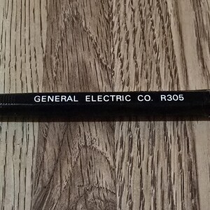 May include: A black mechanical pencil with the text "GENERAL ELECTRIC CO. R305" printed in white. The pencil has a silver tip and a black eraser cap. The pencil is lying on a wooden surface.