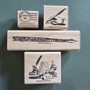 May include: A collection of wooden stamps with black ink designs. The stamps include a New York postmark, a hand holding a quill, a detailed pen, and a scene with books, a quill, and a letter. The text "2000 STAMPIN' UP!" is printed on each stamp.