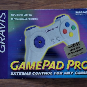 Pode incluir: Uma caixa Gravis Gamepad Pro. A caixa é principalmente azul com um efeito gradiente. O gamepad é branco com botões coloridos. O texto na caixa inclui "100% DIGITAL CONTROL", "10 PROGRAMMABLE BUTTONS" e "EXTREME CONTROL FOR ANY GAME!"