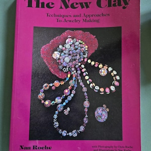 Könnte beinhalten: Ein Buch mit dem Titel "The New Clay" mit einem magentafarbenen Einband. Das Cover zeigt ein farbenfrohes Schmuckdesign aus Perlen und Ton. Der Untertitel des Buches lautet "Techniques and Approaches To Jewelry Making". Autor ist Nan Roche.